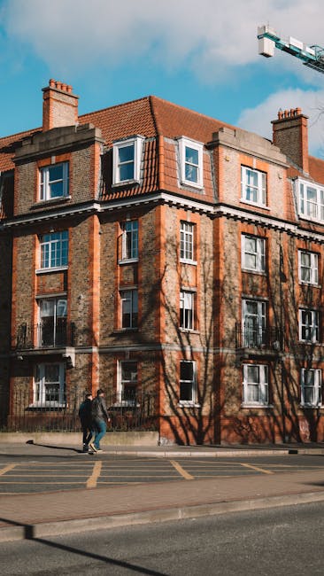 A multi-storey red-brick building with white-framed windows and a sloped red tiled roof, situated along a city street on a clear day with blue sky and some clouds. Shadows of leafless trees are cast onto the building's facade. In front of the building, two movers from Removal Van Fulham are unloading cardboard boxes wrapped in plastic and fabric blankets from a large white moving van parked on the pavement. The movers are carrying the boxes toward the building entrance, which is partially visible. The area includes a sidewalk and a road with yellow road markings, indicating an urban residential environment suitable for house relocations and furniture transport services.