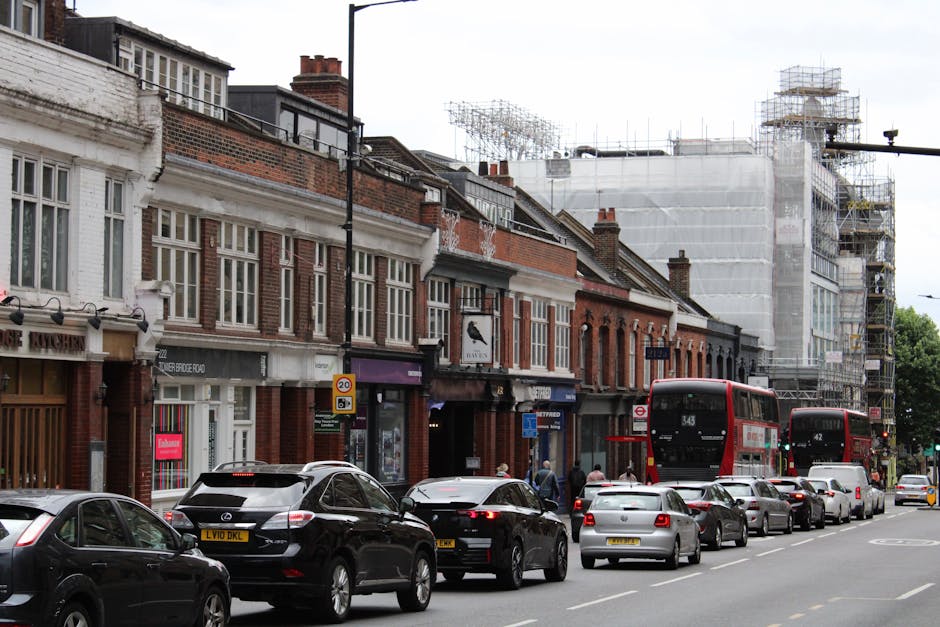 A row of traditional brick and stucco commercial buildings on a busy city street with parked cars and two red double-decker buses in motion. The pavement is lined with several pedestrians, some walking past shopfronts and others entering or leaving the stores. Visible storefronts display signage for various businesses, and the street features traffic signs, including a 20 mph speed limit. In the background, construction scaffolding and a building under renovation are covered with white protective sheeting, indicating ongoing building work. Cars are parked along the curb, while others are in motion, suggesting an active urban environment consistent with house and commercial premises in Fulham. The scene captures typical city logistics involved in home relocation or furniture transport, with a focus on urban loading and moving activities that might require services such as those offered by Removal Van Fulham, as part of a comprehensive removals or moving process.