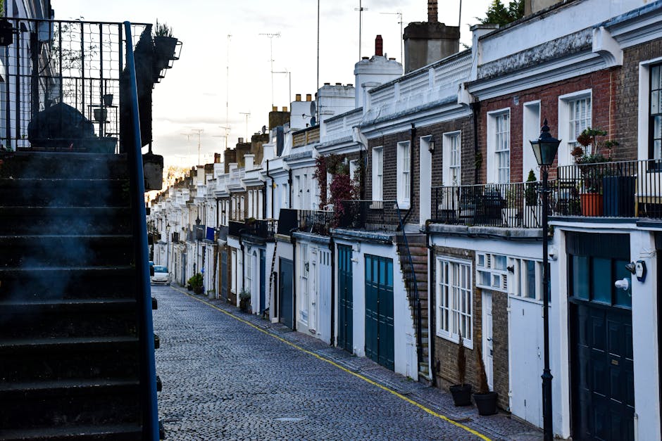 A narrow residential street in Fulham with a row of terraced houses featuring brick facades, white window frames, and small balconies with black metal railings. Several potted plants are placed on the balconies and terraces. The street is cobbled and lined with street lamps mounted on the exterior walls. On the left side of the image, a dark external staircase with a metal railing leads up to an upper level of one of the buildings. The lighting suggests early morning or late afternoon, with soft sunlight casting gentle shadows. This scene illustrates the typical tight urban access environment where house removals and furniture transport require careful planning, and professional moving services like those offered by [COMPANY_NAME] are often needed for smooth home relocation, especially on narrow streets with limited access.