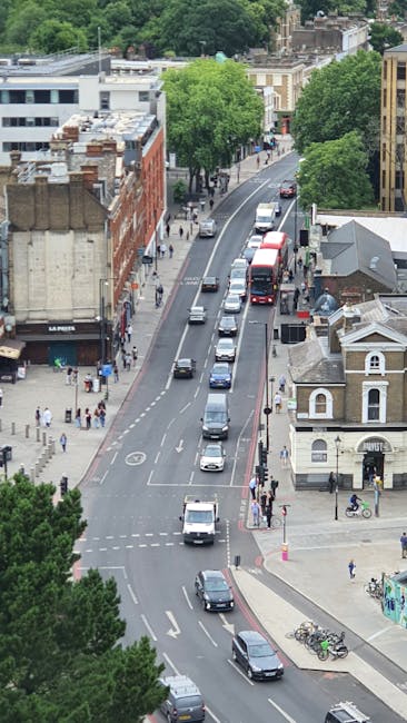 An aerial view of a busy urban street with vehicles including cars, buses, and vans lined up in traffic. Pedestrians are walking on the sidewalks, and bicycles are parked near the curb. Green leafy tr