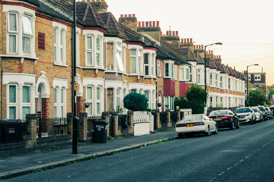 A row of terraced houses in Sands End, Fulham, featuring brick exteriors with white-framed bay windows and decorative cornices. The pavement in front of the houses is lined with small gardens enclosed by low brick walls and iron railings. Parked along the street are various cars, including sedans and hatchbacks, some partially obscured by trees and lampposts. A black wheelie bin is visible on the sidewalk near one of the houses. The scene is lit by natural daylight, with a soft sunset glow in the background, indicating early evening. The image captures the quiet residential area typical for home relocations and furniture transport services, with front yards prepared for packing and loading activities, suggesting ongoing or upcoming house removals facilitated by [COMPANY_NAME].