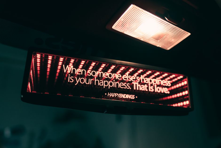 Close-up view of an indoor ceiling area showing a rectangular LED digital display panel with red and white text reading 'When someone else's happiness is your happiness. That is love,' mounted beneath a modern ceiling light fixture with a transparent cover. The display is positioned in a space that appears to be part of a building interior, possibly an office or common area, with the panel supported by a black frame. The ceiling is dark, and the lighting creates a contrast that highlights the digital message. While the image does not directly depict furniture or moving activities, it suggests an indoor environment where lighting and visual messaging are present, aligning with themes of interior spaces often involved in house removals or home relocation preparations, as managed by companies like Removal Van Fulham.